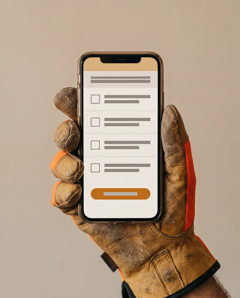 Photograph of a worn work glove holding a phone displaying a per-pay-period attestation card — four checkbox rows with text shown as plain grey bars and the confirm control highlighted in amber.